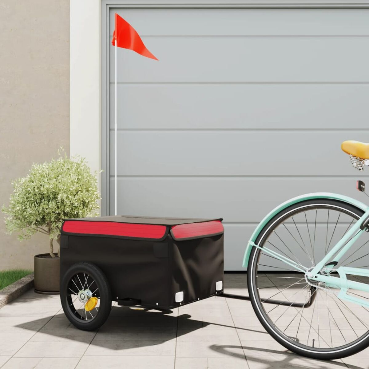 Ein schwarzer Anhänger an einem blauen Fahrrad mit roter Flagge. Garage und Busch im Hintergrund.