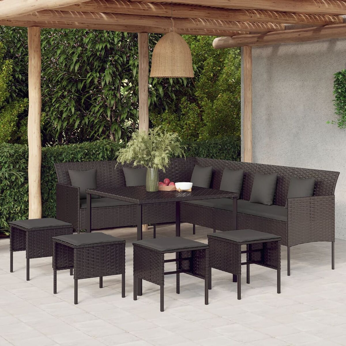Terrassenset mit Tisch, Bänken und Hockern. Dunkelbraunes Korbgeflecht mit grauen Kissen. Grünes Laub im Hintergrund.