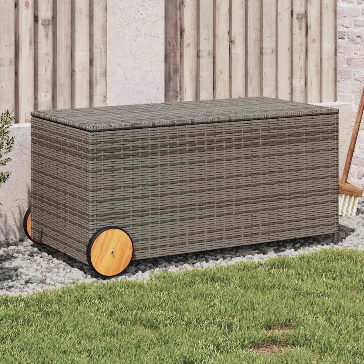 Truhe im Rattan-Stil mit Rädern, auf einer Stein- und Grasfläche, in der Nähe eines Holzzauns.