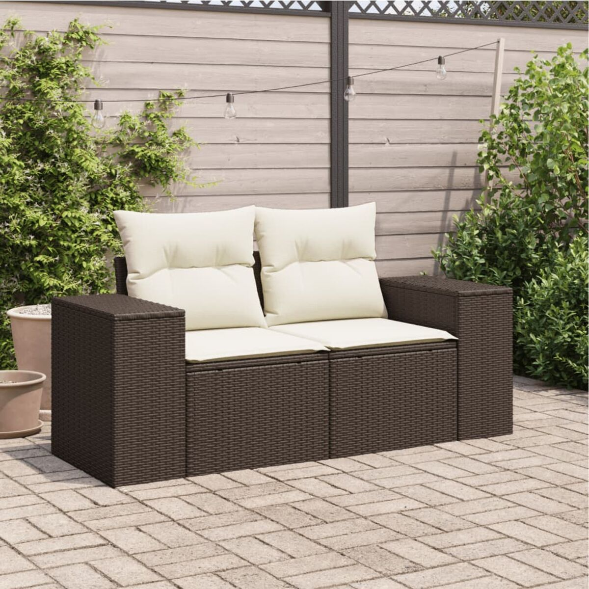 Braunes Outdoor-Sofa aus Korbgeflecht mit weißen Kissen auf gepflasterter Terrasse, Pflanzen im Hintergrund.