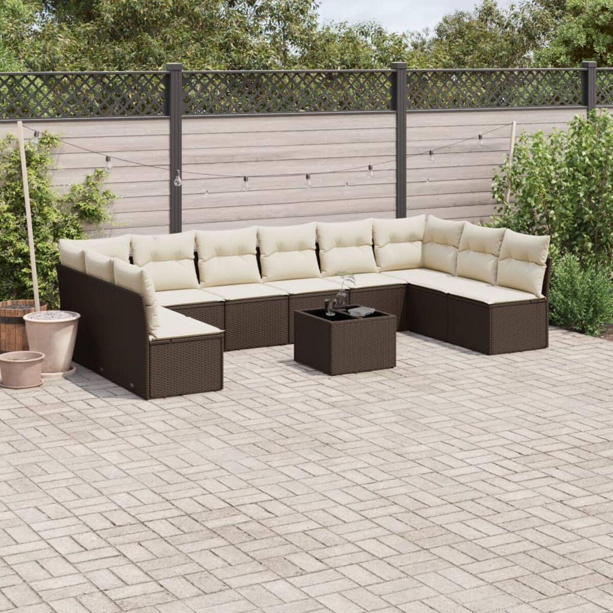 Divano da esterno in rattan marrone con cuscini color crema e tavolino quadrato. Recinzione e piante sullo sfondo.