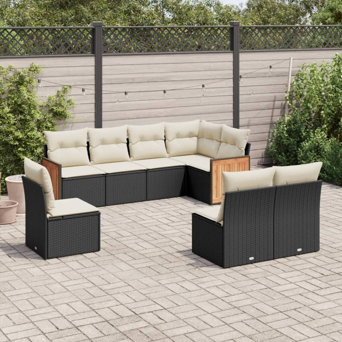 Schwarzes Ecksofa aus Korbgeflecht mit cremefarbenen Kissen. Holzarmlehnen. Gepflasterte Terrasse, Holzzaun im Hintergrund.