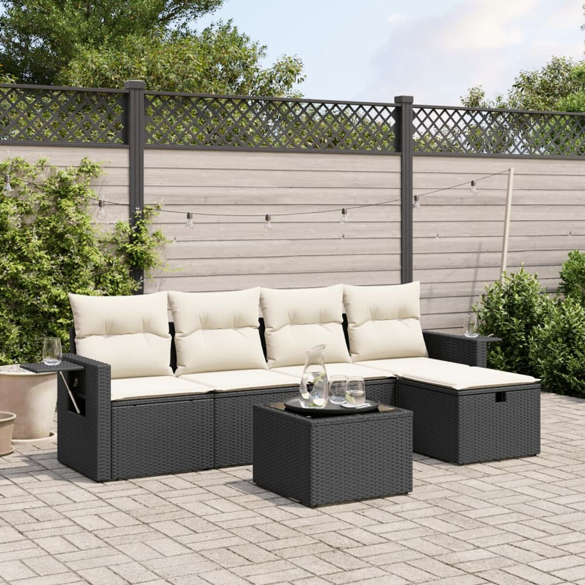 Ein schwarzes Korbmöbel-Sofa mit cremefarbenen Kissen, einem Tisch und einer Backsteinterrasse. Ein Zaun ist im Hintergrund.