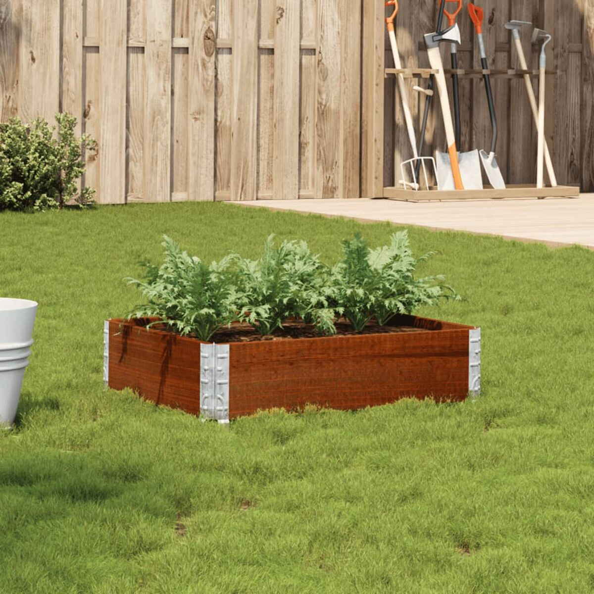 Fioriera in legno con piante su erba verde con attrezzi da giardinaggio sullo sfondo.