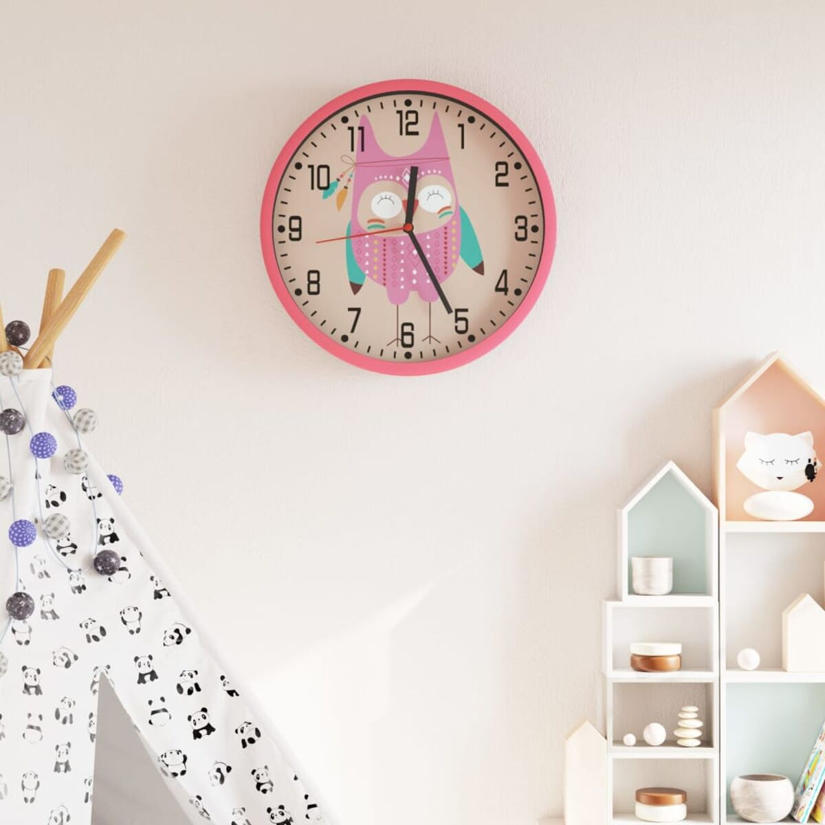 Eine rosa Eulen-Uhr an einer weißen Wand. Im Bild sind ein Tipi und Regale mit Dekorationen zu sehen.