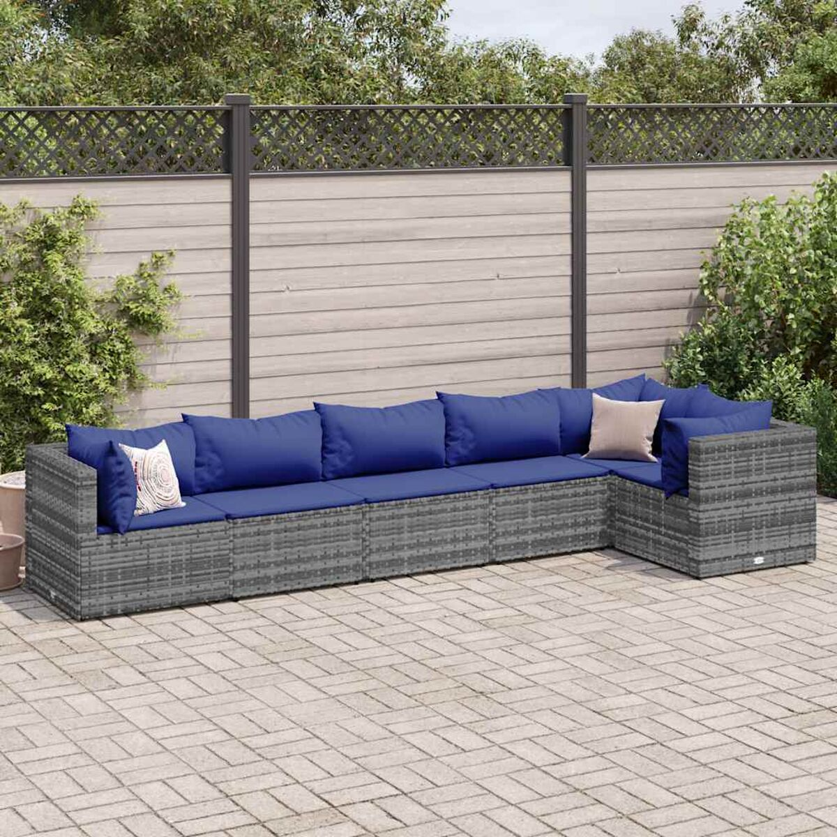 Divano a L in rattan grigio con cuscini blu. Su un patio in mattoni, con recinzione e verde.