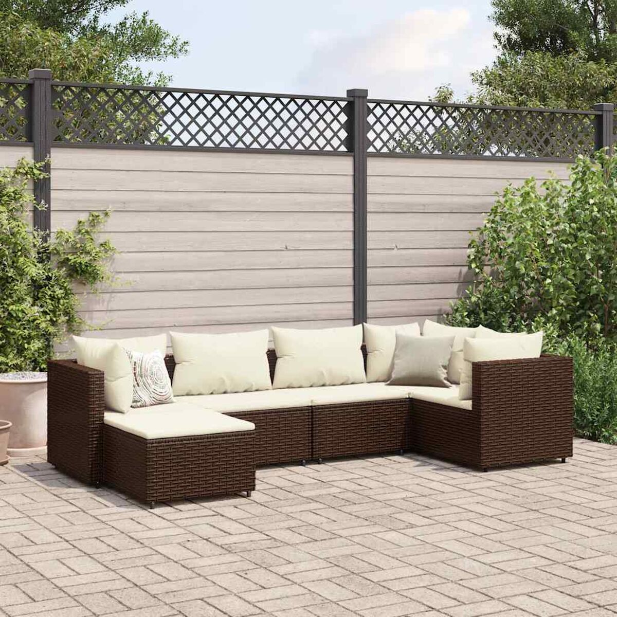 Divano componibile in vimini marrone con cuscini color crema, su un patio in mattoni. Recinzione e verde sullo sfondo.