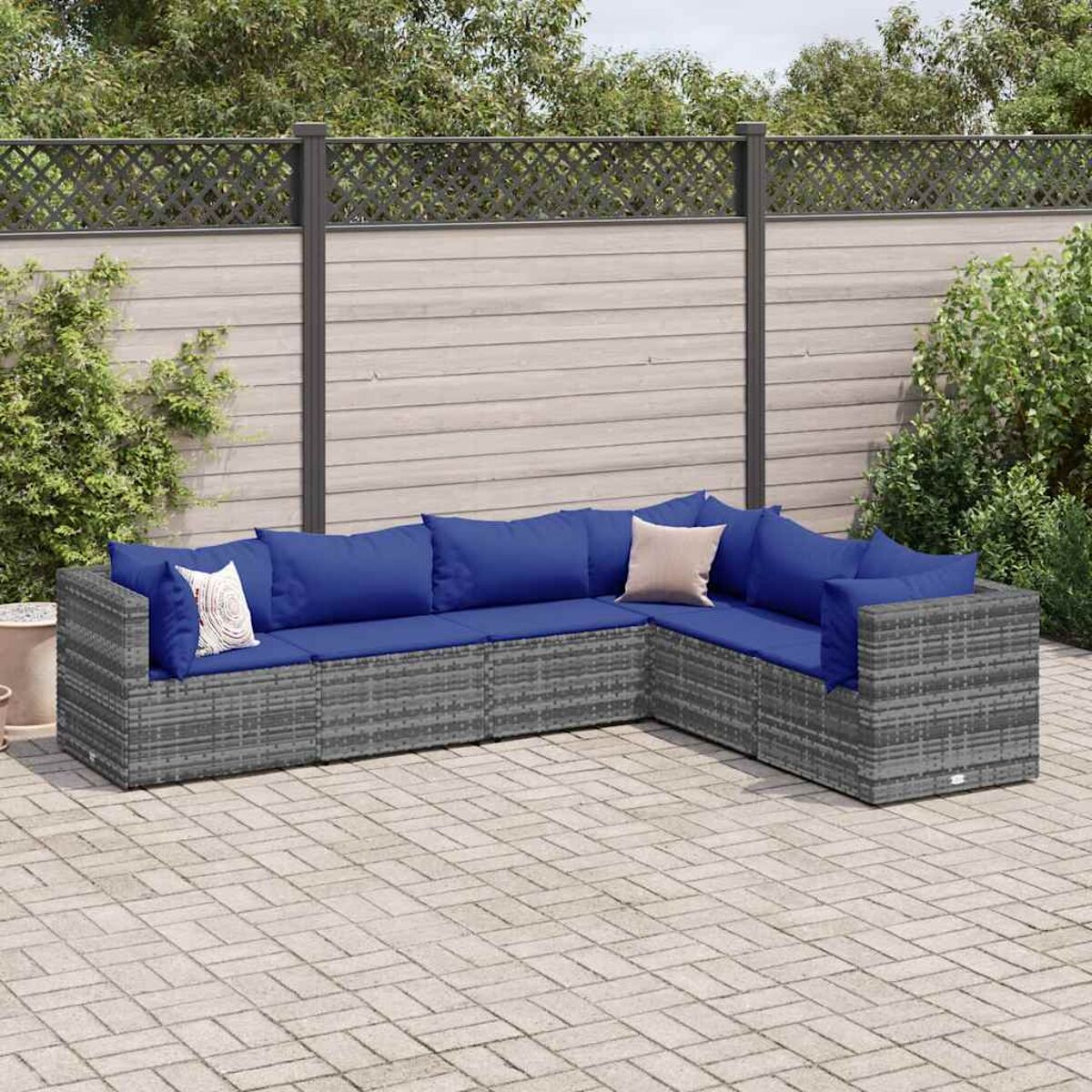 Divano angolare in rattan grigio con cuscini blu. Su un patio pavimentato, con recinzione in legno.