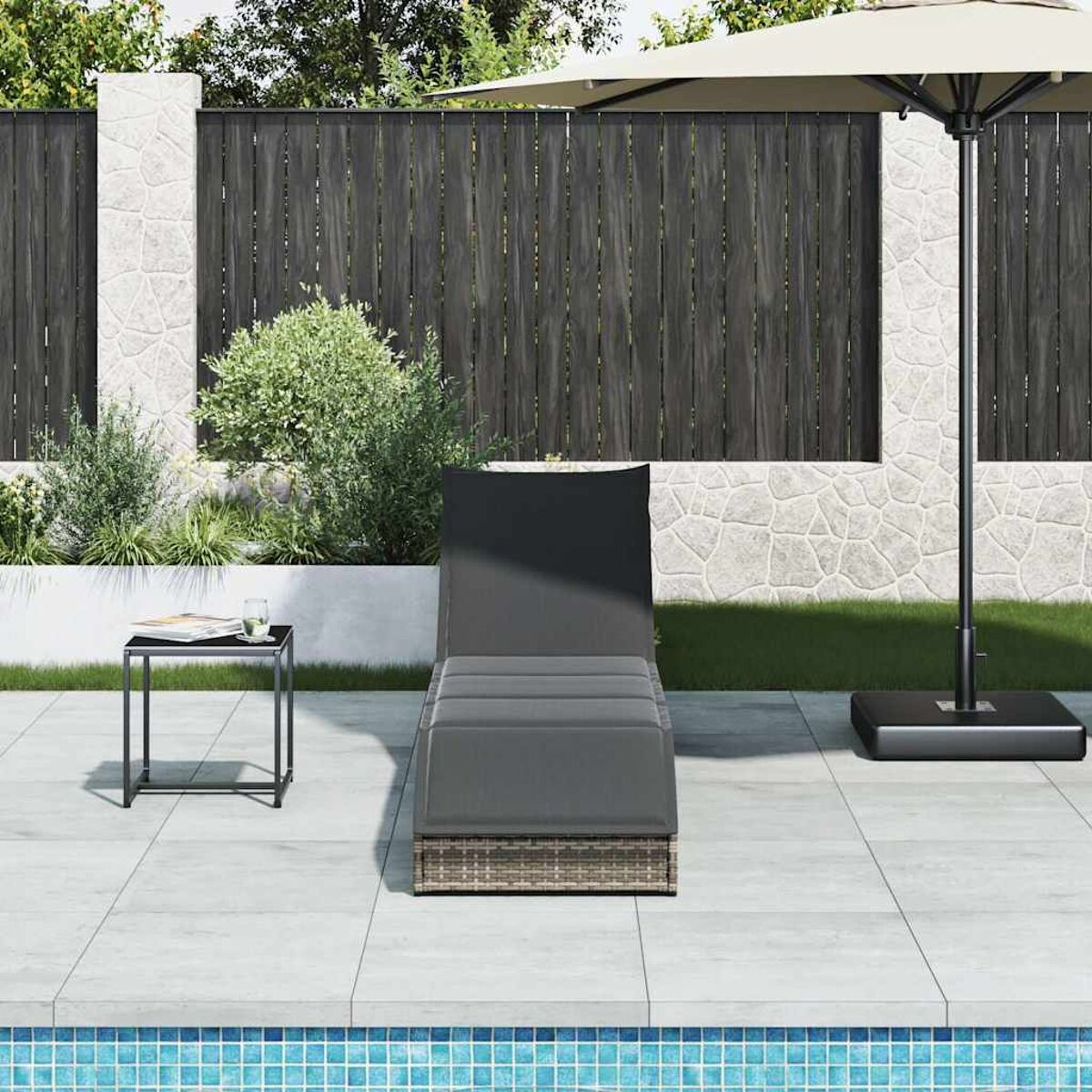 Una chaise longue grigia con un piccolo tavolino nero e un ombrellone vicino a una piscina, contro una recinzione.