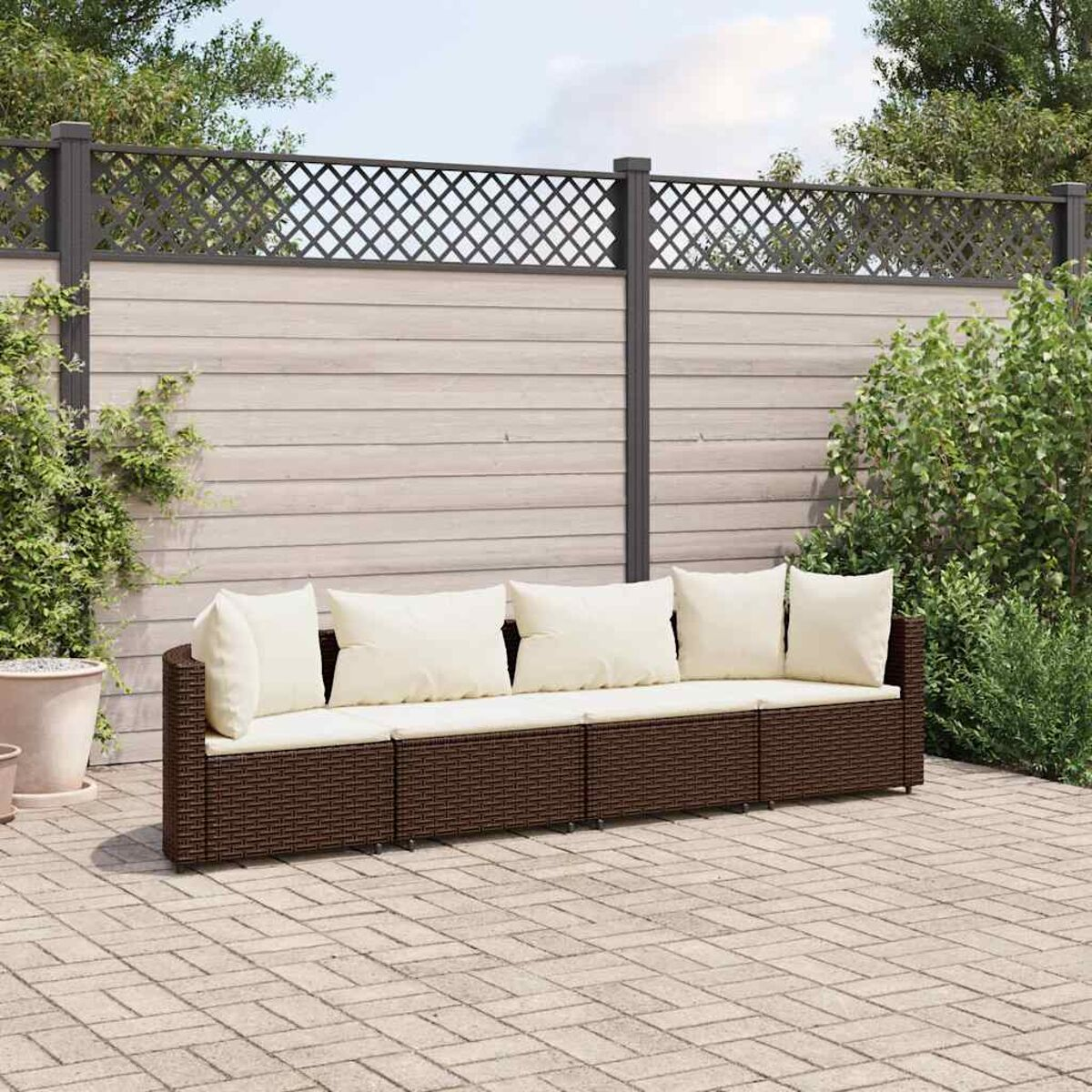 Divano da esterno in vimini marrone con cuscini color crema. Recinzione in legno e piante sullo sfondo. Scena all'aperto soleggiata.