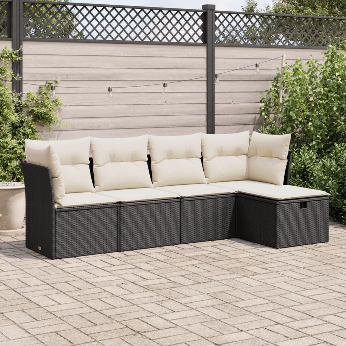 Divano a L in vimini grigio scuro con cuscini color crema su un patio in mattoni, con una recinzione sullo sfondo.