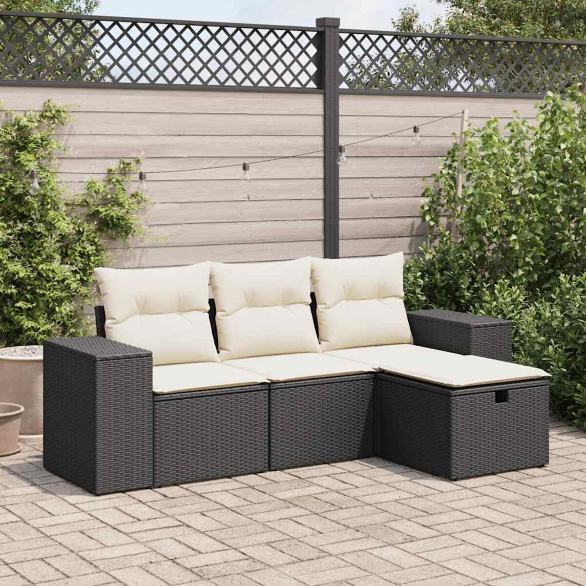 Dunkelgraues Rattan-Outdoor-Sofa mit weißen Kissen auf einer Ziegelterrasse. Grüne Pflanzen und Zaun im Hintergrund.