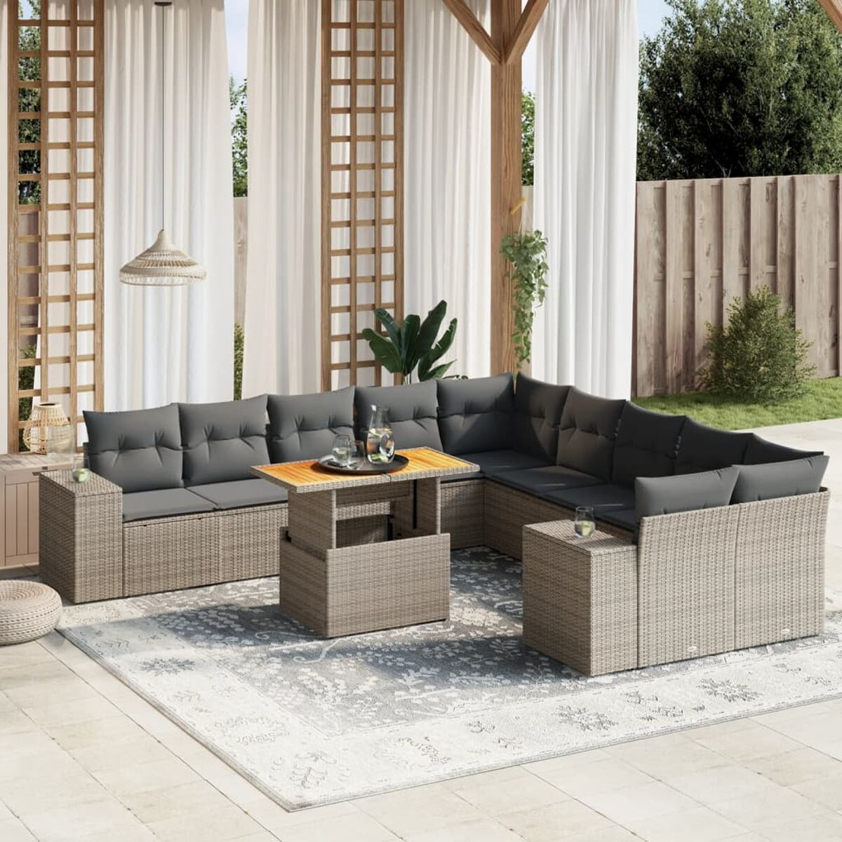 Graues Outdoor-Wickermöbel-Set mit Kissen, Tisch und Teppich. Unter einer Pergola, mit Vorhängen und Pflanzen.