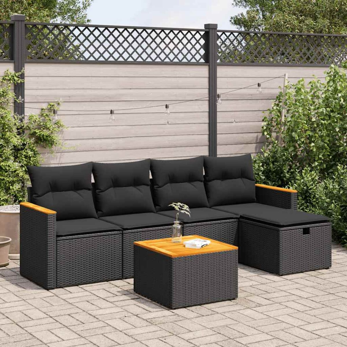 Geflochtenes Outdoor-Sofa-Set. Schwarze Kissen, dunkelgraues Rattan. Tisch davor. Hintergrund: Zaun und Pflanzen.