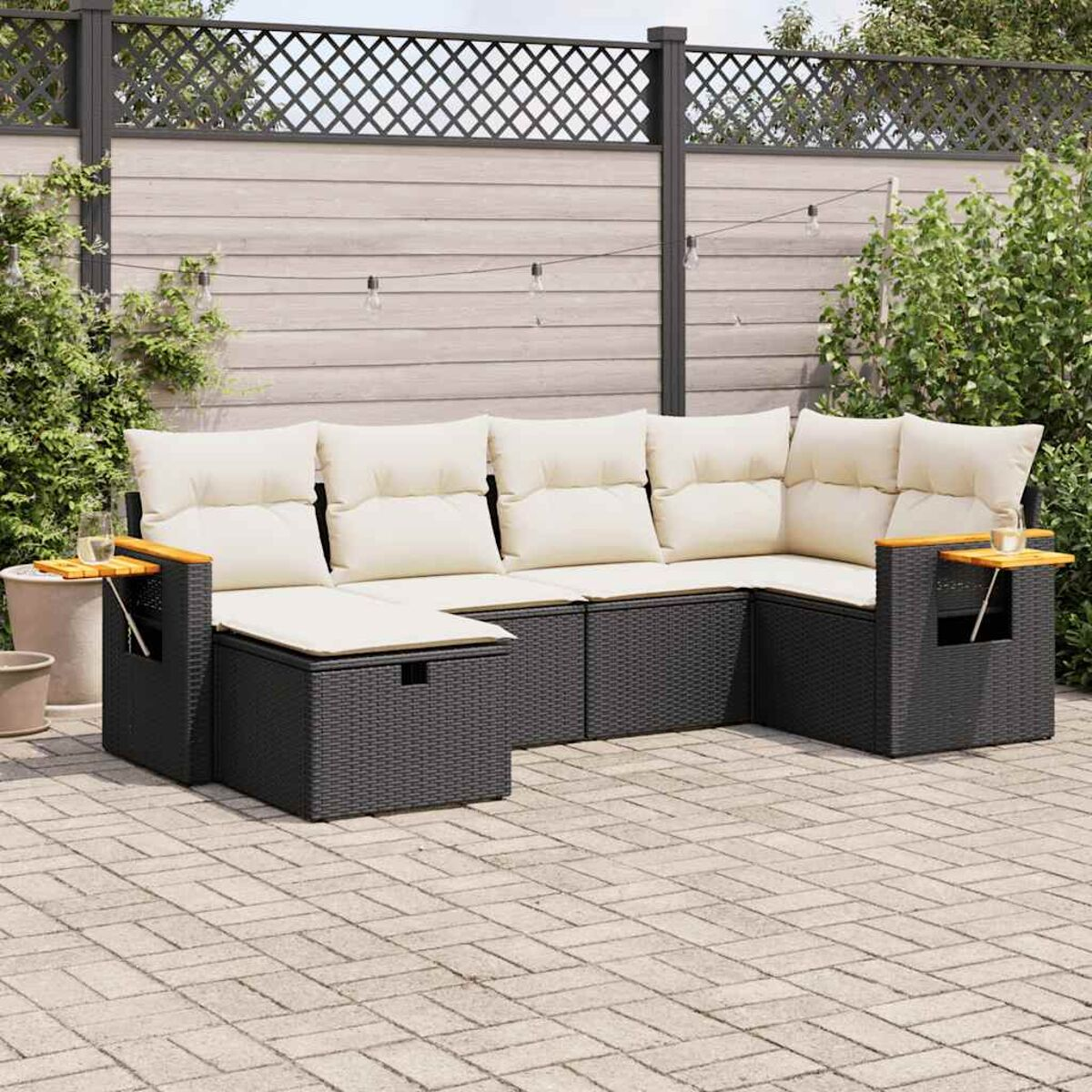 Schwarzes Outdoor-Ecksofa mit cremefarbenen Kissen. Es hat Beistelltische. Gepflasterte Terrasse, grüner Hintergrund.