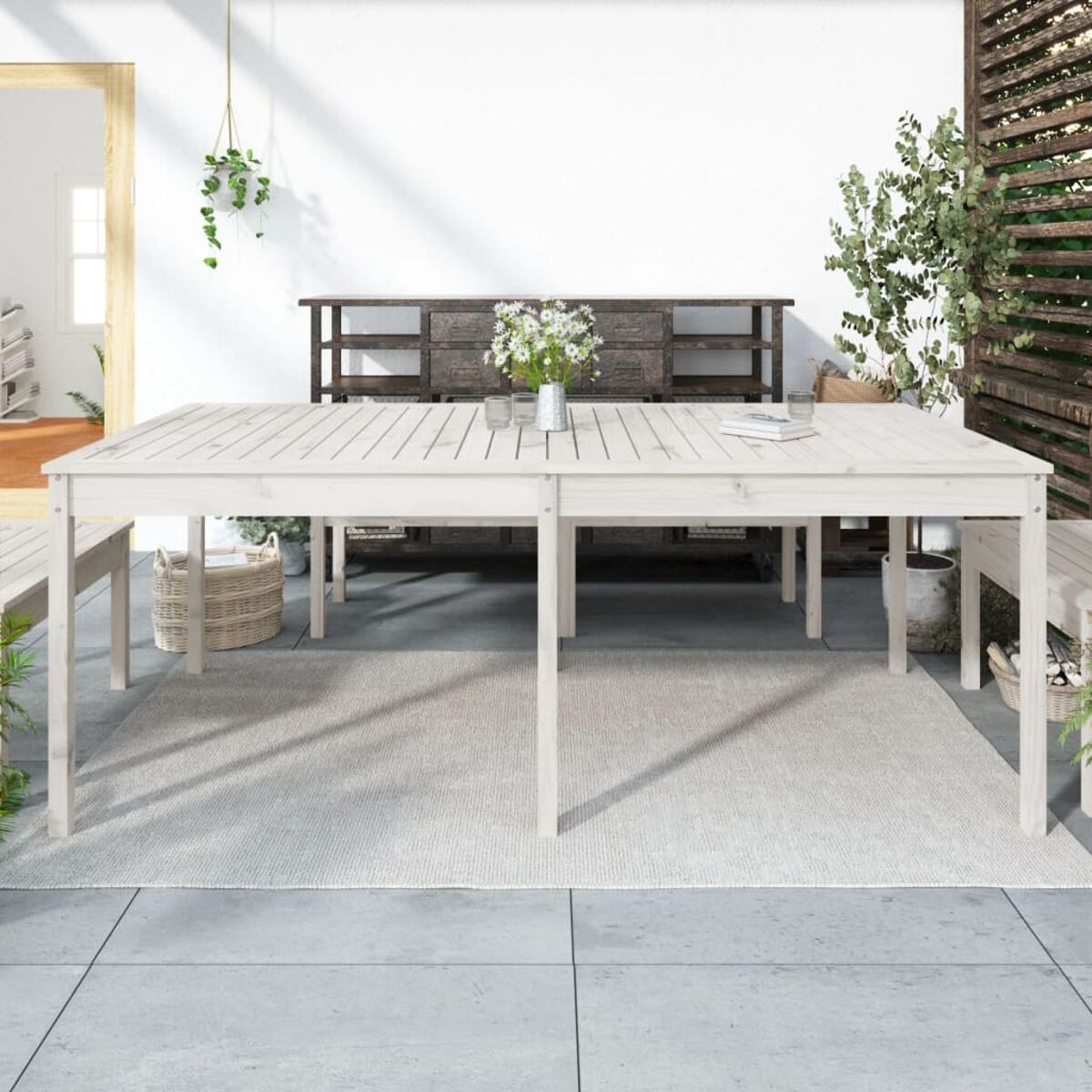 Weißer Holztisch im Freien mit hellem Teppich. Blumen, Pflanzen und ein Holzschrank sind vorhanden.