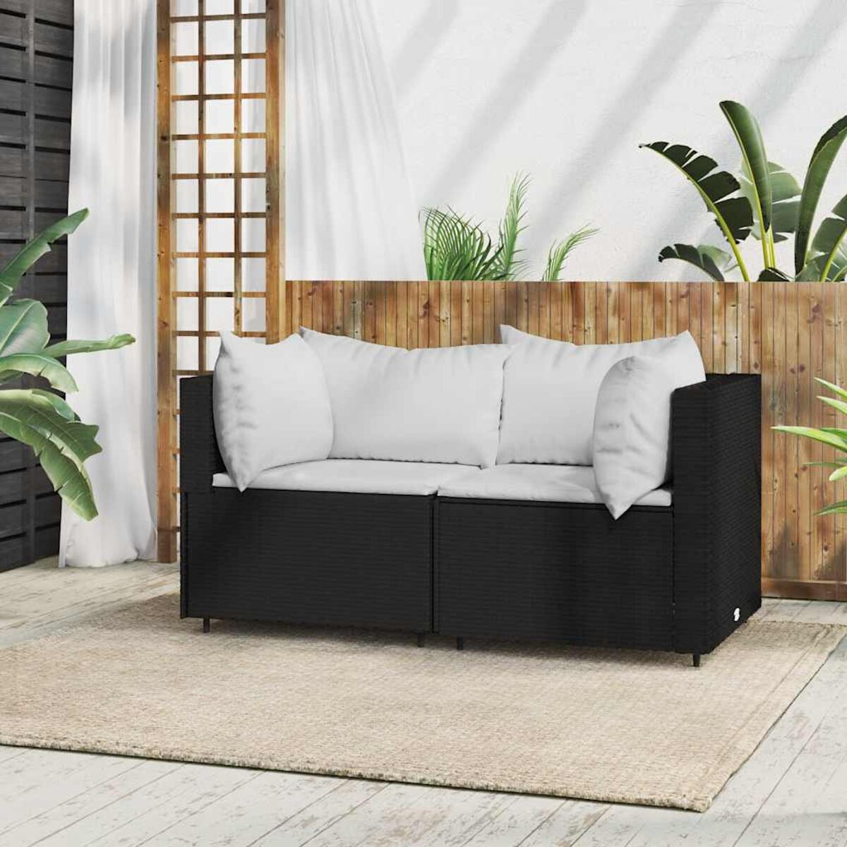 Ein schwarzes Outdoor-Sofa mit weißen Kissen, auf einem beigen Teppich, vor einem Holzzaun.