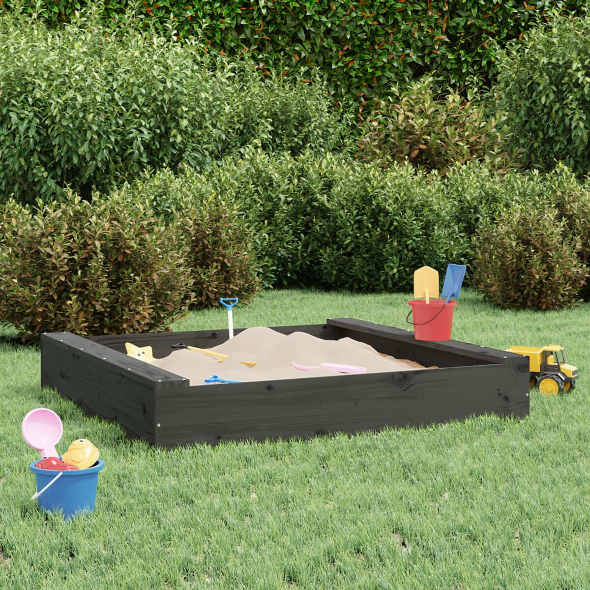 Schwarzer Sandkasten mit Spielzeug auf Gras, umgeben von Büschen und Mauer.