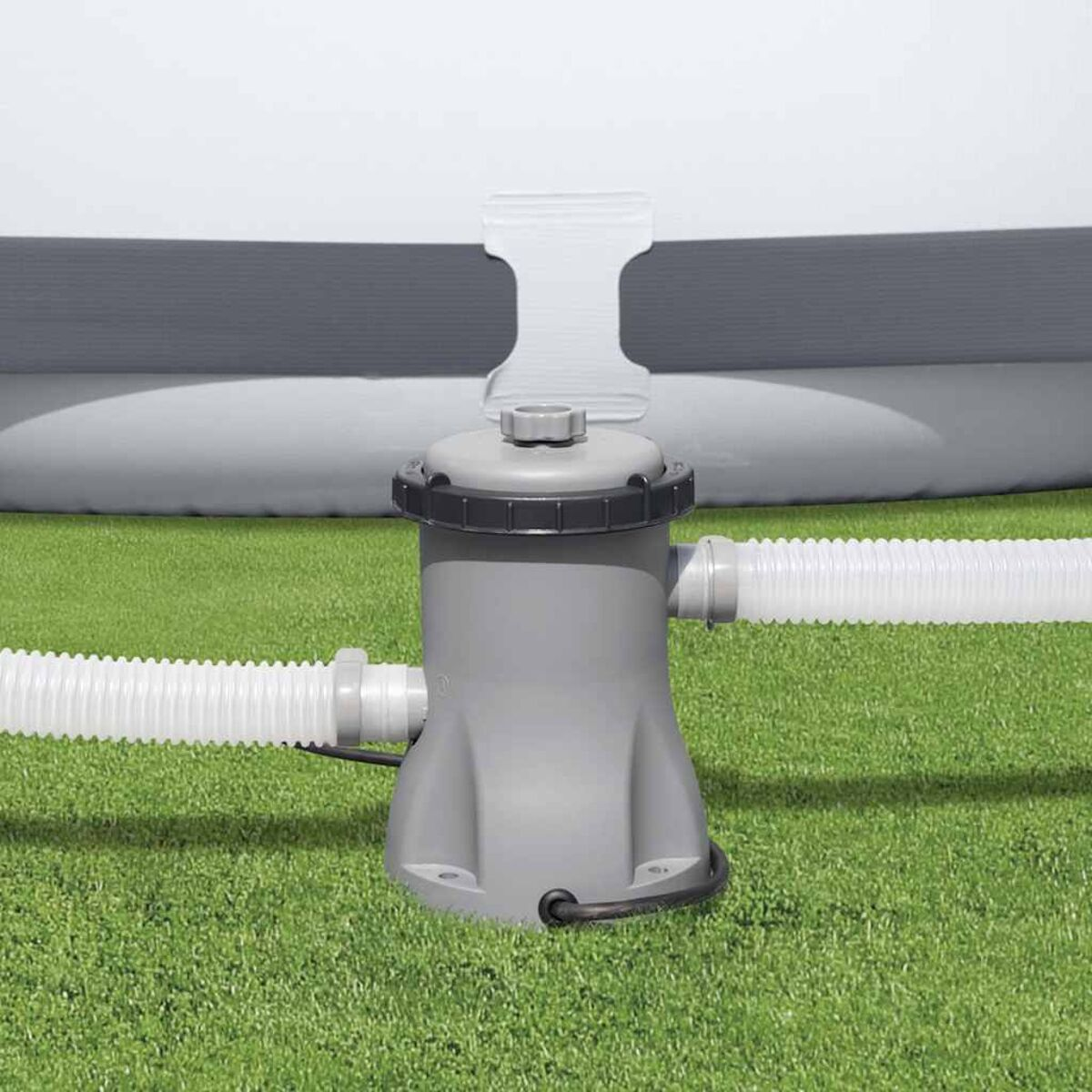 Eine graue Poolpumpe mit zwei weißen Schläuchen in einem grünen Grasbereich.
