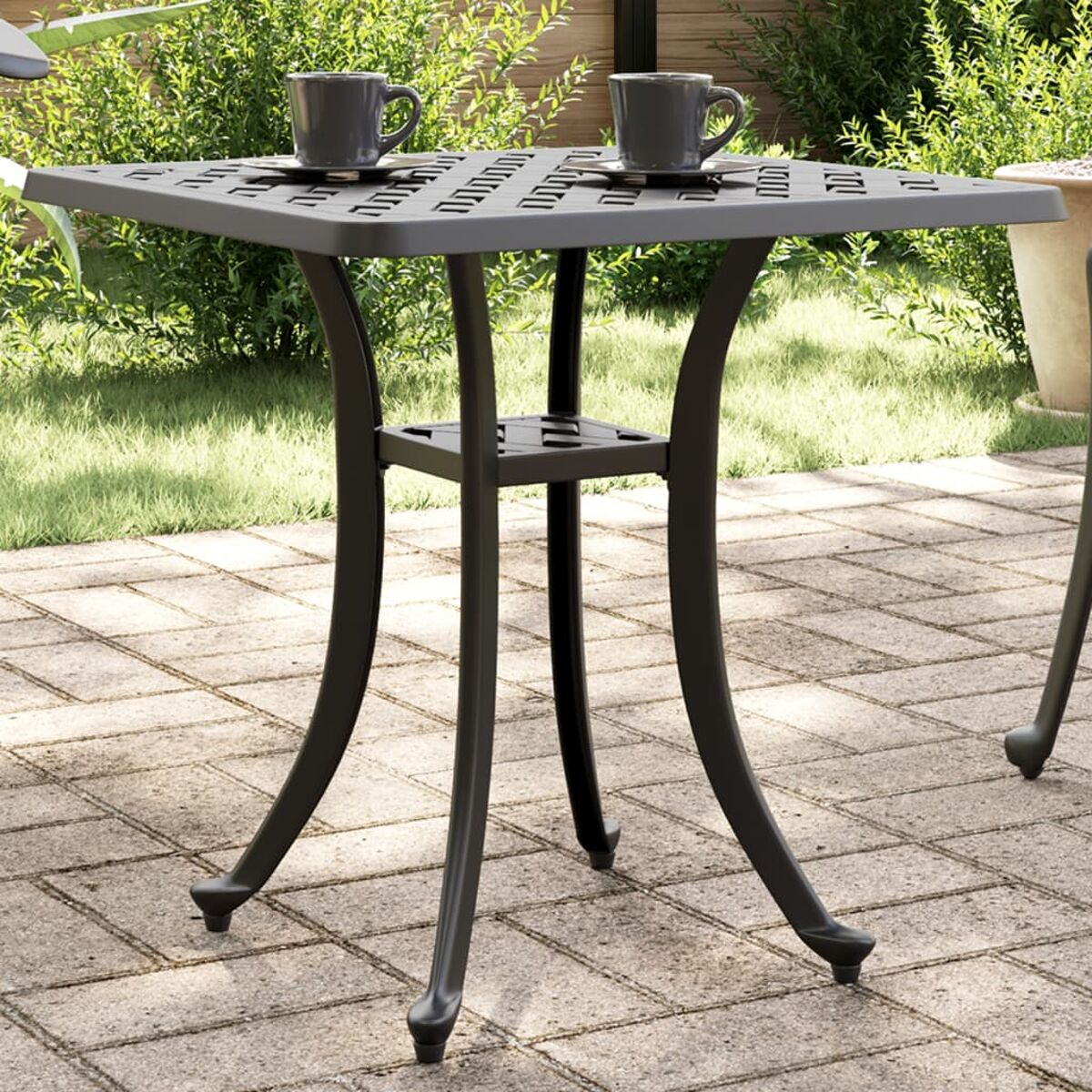 Schwarzer Metall-Terrassentisch mit zwei Tassen. Der Tisch steht auf einer Ziegelpflasterung, im Hintergrund Grün.