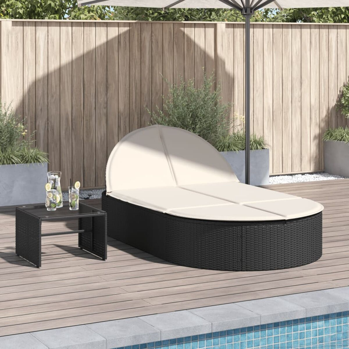 Eine schwarze Wicker-Chaiselongue mit weißem Kissen steht auf einem Holzdeck. Ein kleiner Tisch und ein Sonnenschirm vervollständigen die Szene.