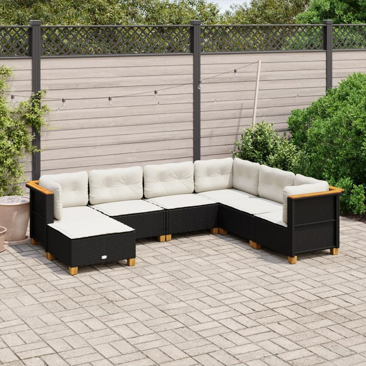 U-förmiges Outdoor-Sofa mit cremefarbenen Kissen. Schwarzer Korbrahmen, Holzarmlehnen. Auf einer Terrasse mit Zaun im Hintergrund.