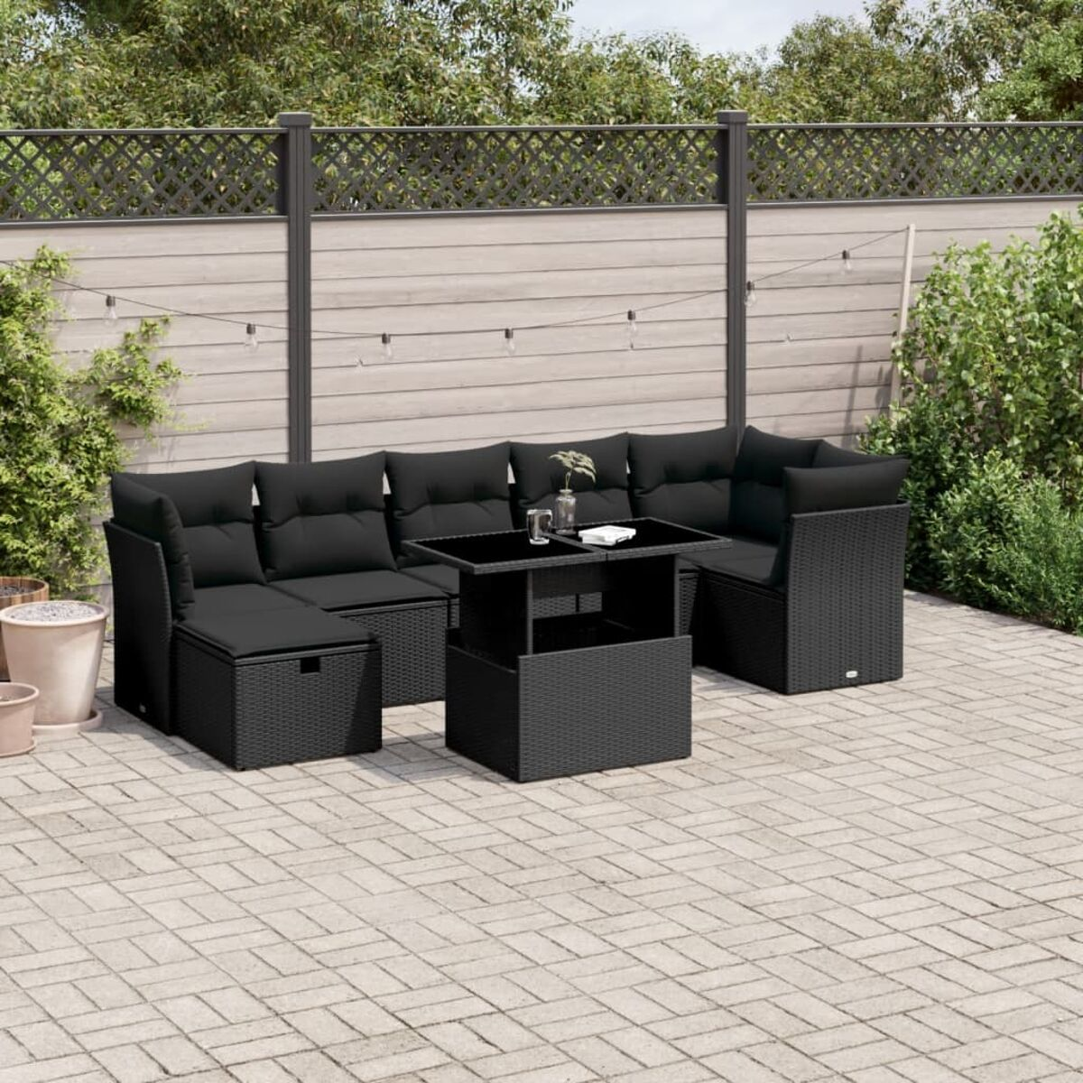 Schwarzes Gartenmöbel-Set aus Korbgeflecht mit schwarzen Kissen und einem schwarzen Tisch. Gepflasterte Terrasse mit Pflanzen.