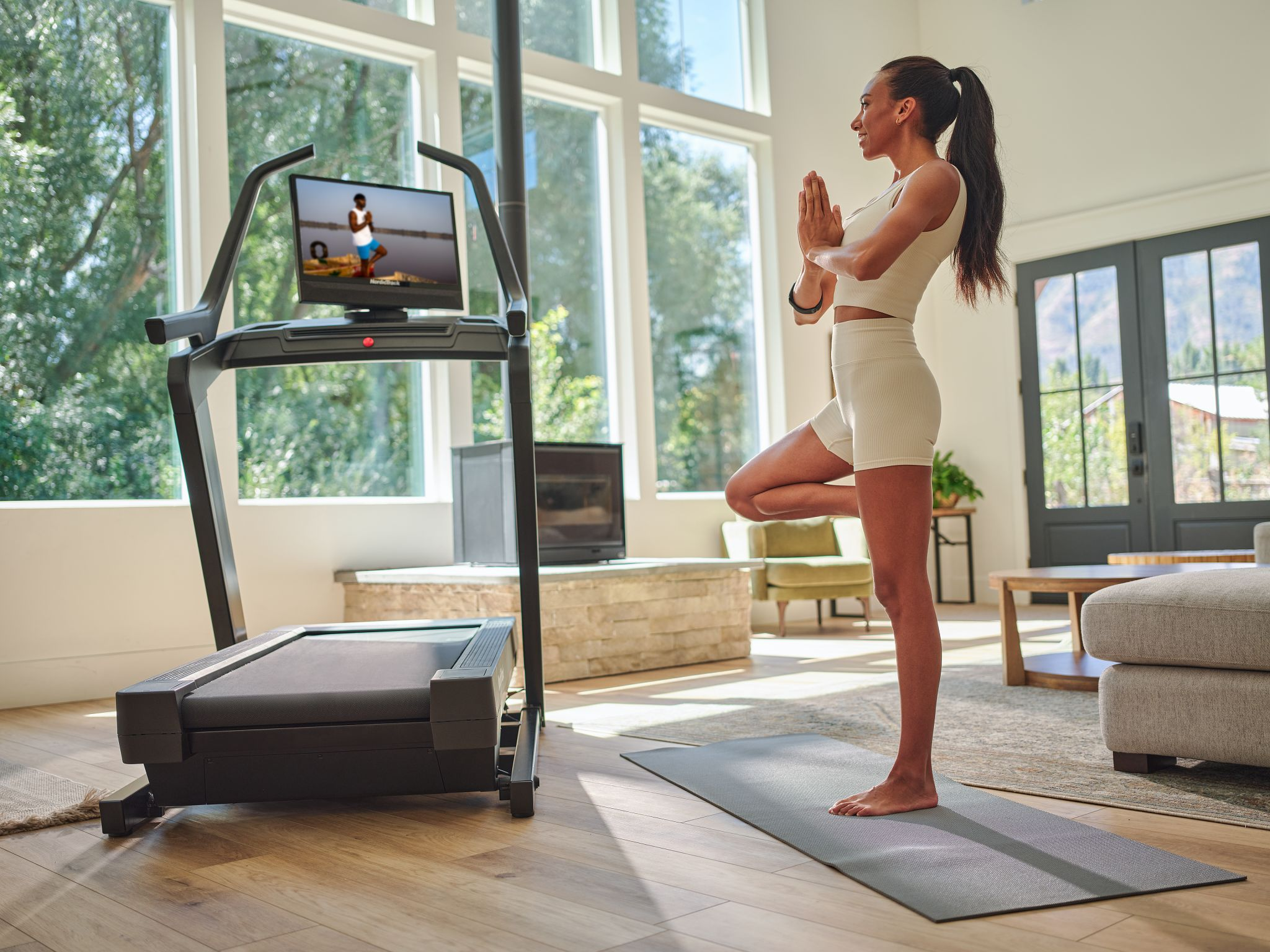 Frau macht Yoga auf einer Matte in der Nähe eines Laufbands, in einem hellen Raum.