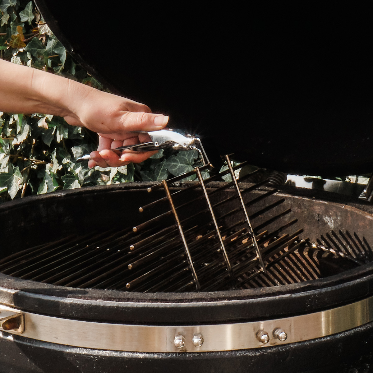 Hand holding a tool cleaning a grill grate. Background: grill and foliage.