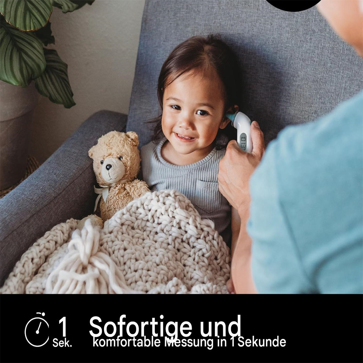 Ein Kleinkind im Stuhl wird die Temperatur gemessen. Ein Teddybär und eine Decke sind in der Nähe.
