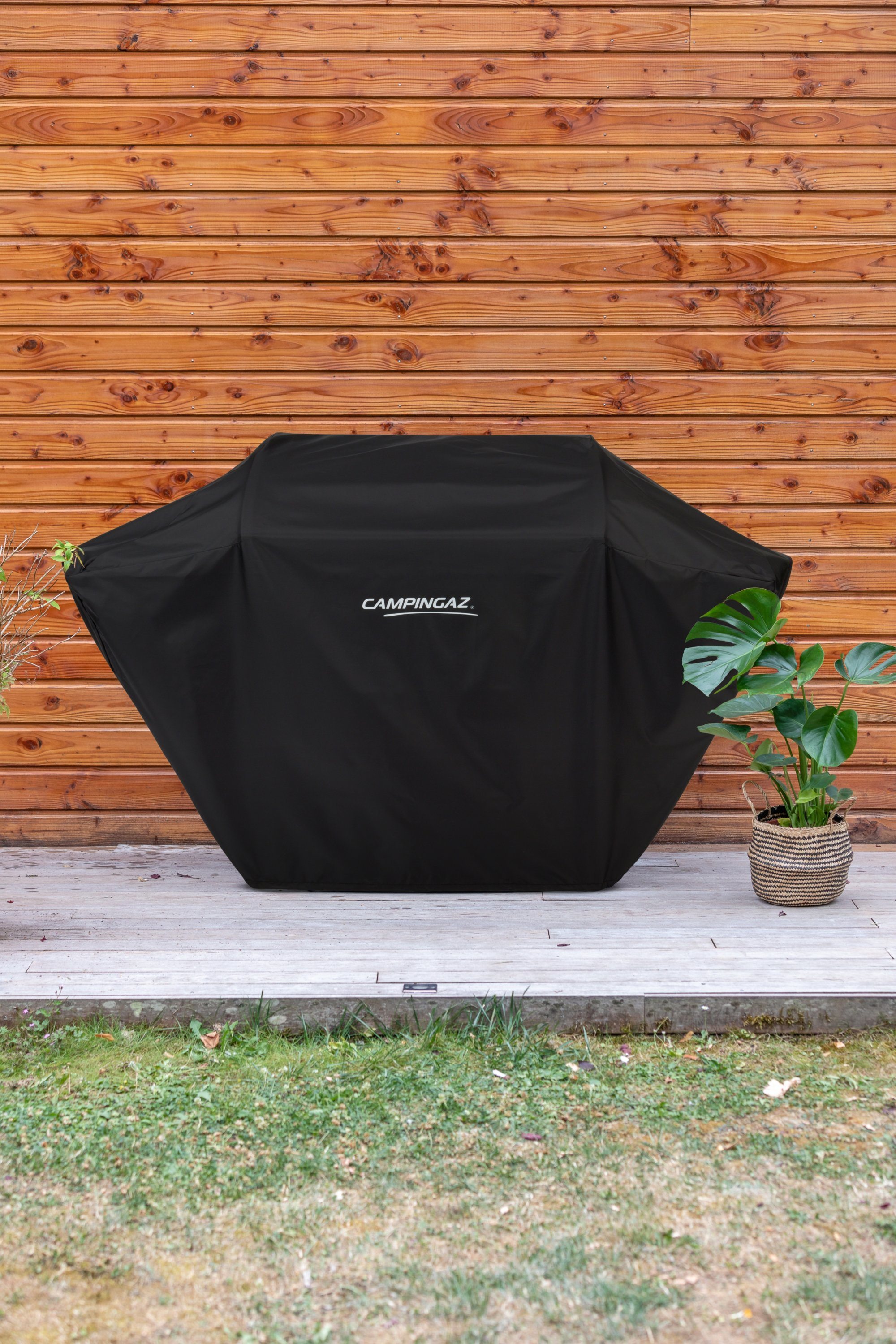 Czarna osłona grilla z białym logo, obok rośliny.