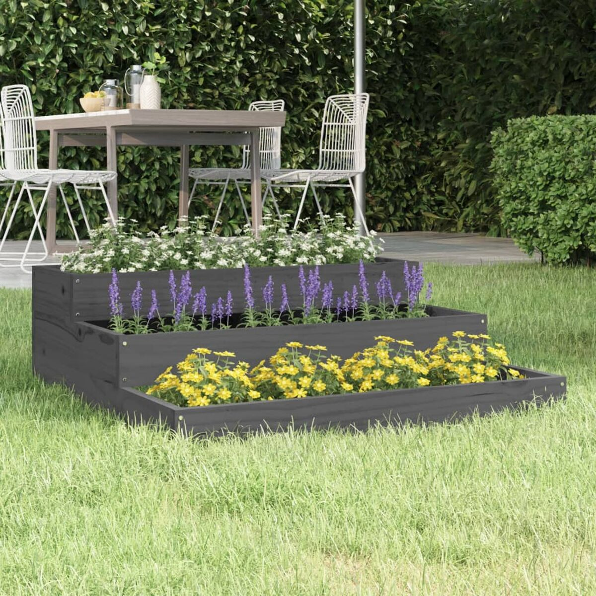 Dreistufiger grauer Pflanzkübel mit Blumen. Tisch und Stühle im Hintergrund.