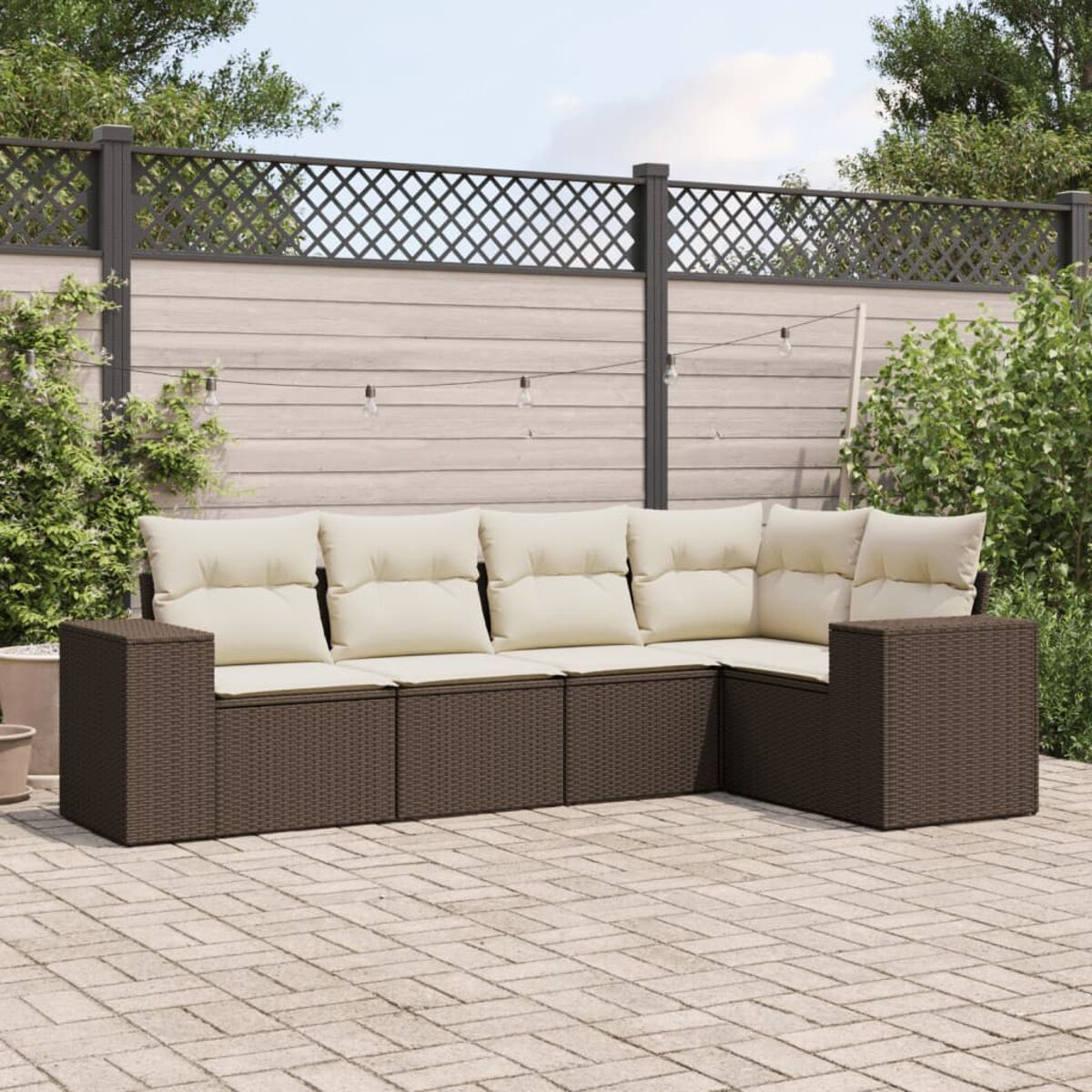 Divano componibile in vimini marrone con cuscini beige su un patio. Recinzione illuminata sullo sfondo.