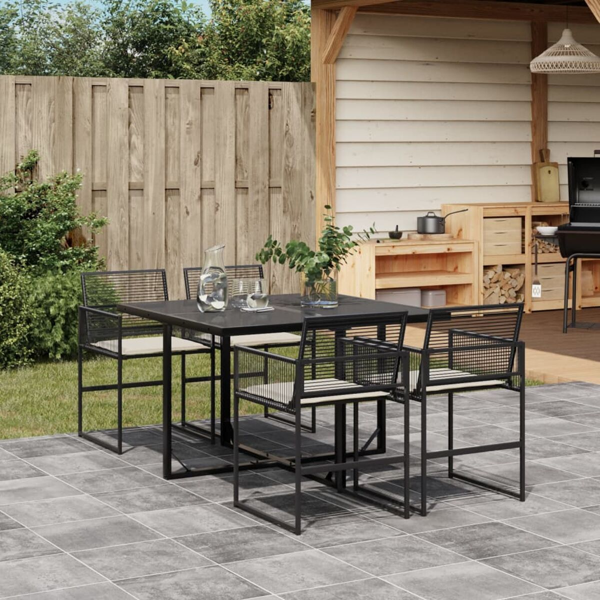 Essgruppe im Freien: schwarzer Metalltisch und Stühle. Tisch mit Glas und Pflanzen, auf gepflasterter Terrasse. Holzschuppen im HG.