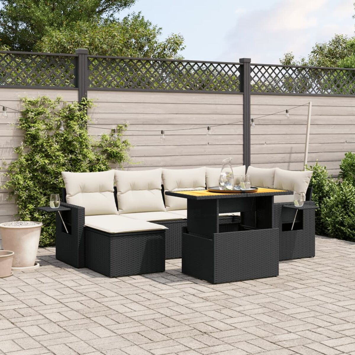 Outdoor-Möbelset aus schwarzem Korbgeflecht, bestehend aus Sofa, Ottomanen und Tisch. Pflastersteine und Pflanzen im Hintergrund.
