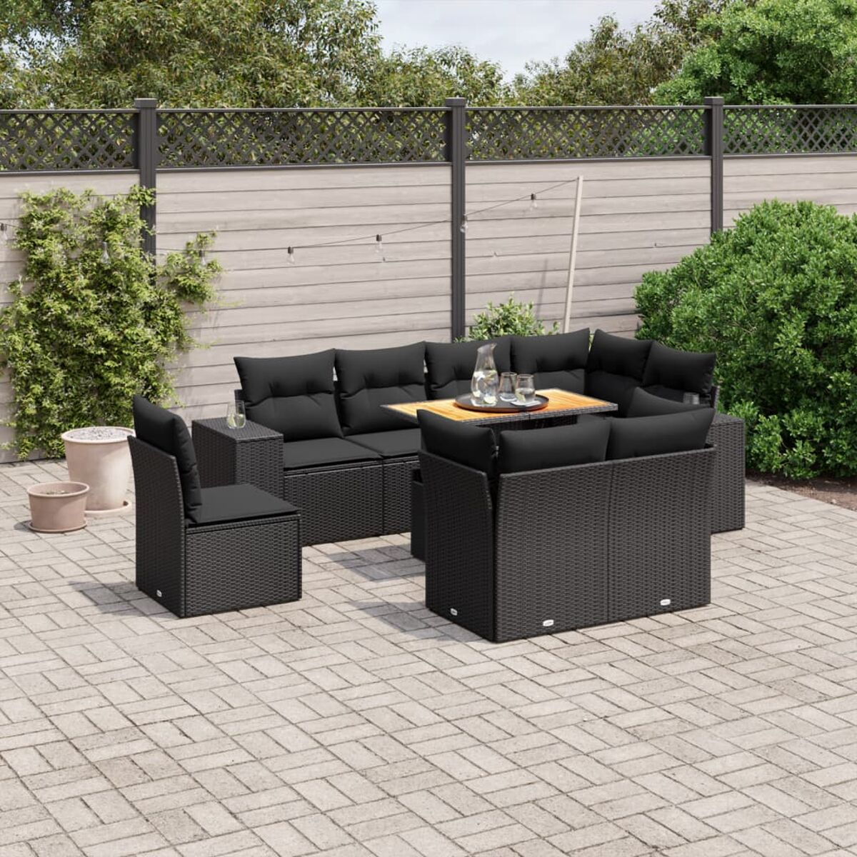 Outdoor-Möbelset aus schwarzem Korbgeflecht, bestehend aus Sofa, Stuhl und Tisch. Pflastersteine und Pflanzen im Hintergrund.