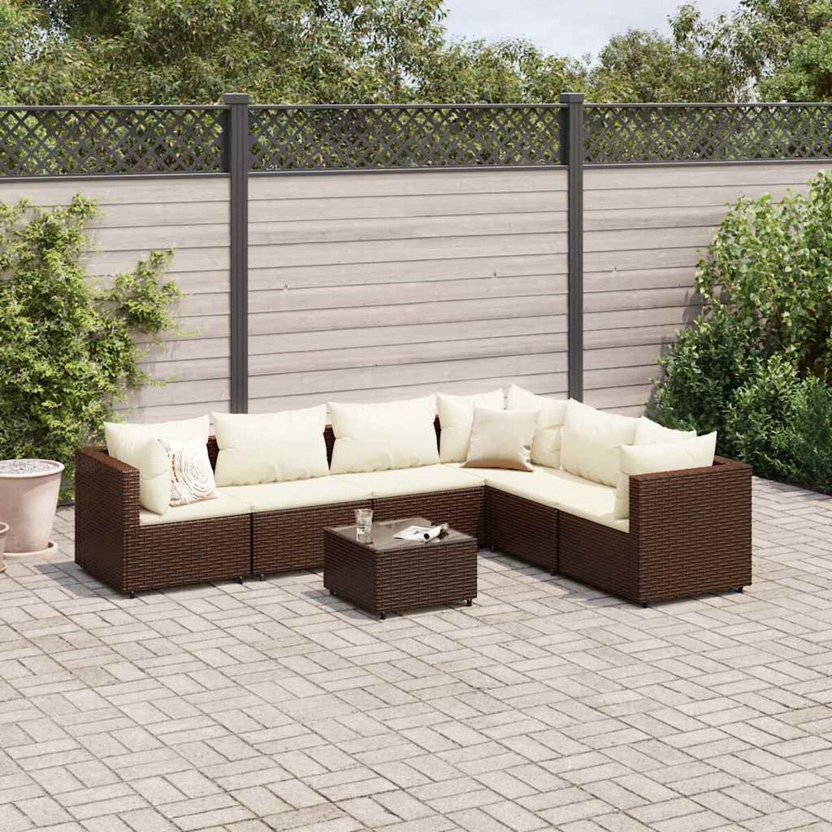 Divano angolare in vimini marrone con cuscini beige, tavolino su un patio in pietra. Recinzione in legno e cespugli verdi sullo sfondo.