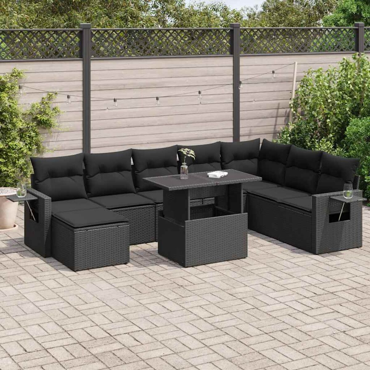 Set divano in vimini nero da esterno con tavolo, su un patio. Include cuscini e tavolini, contro una recinzione.