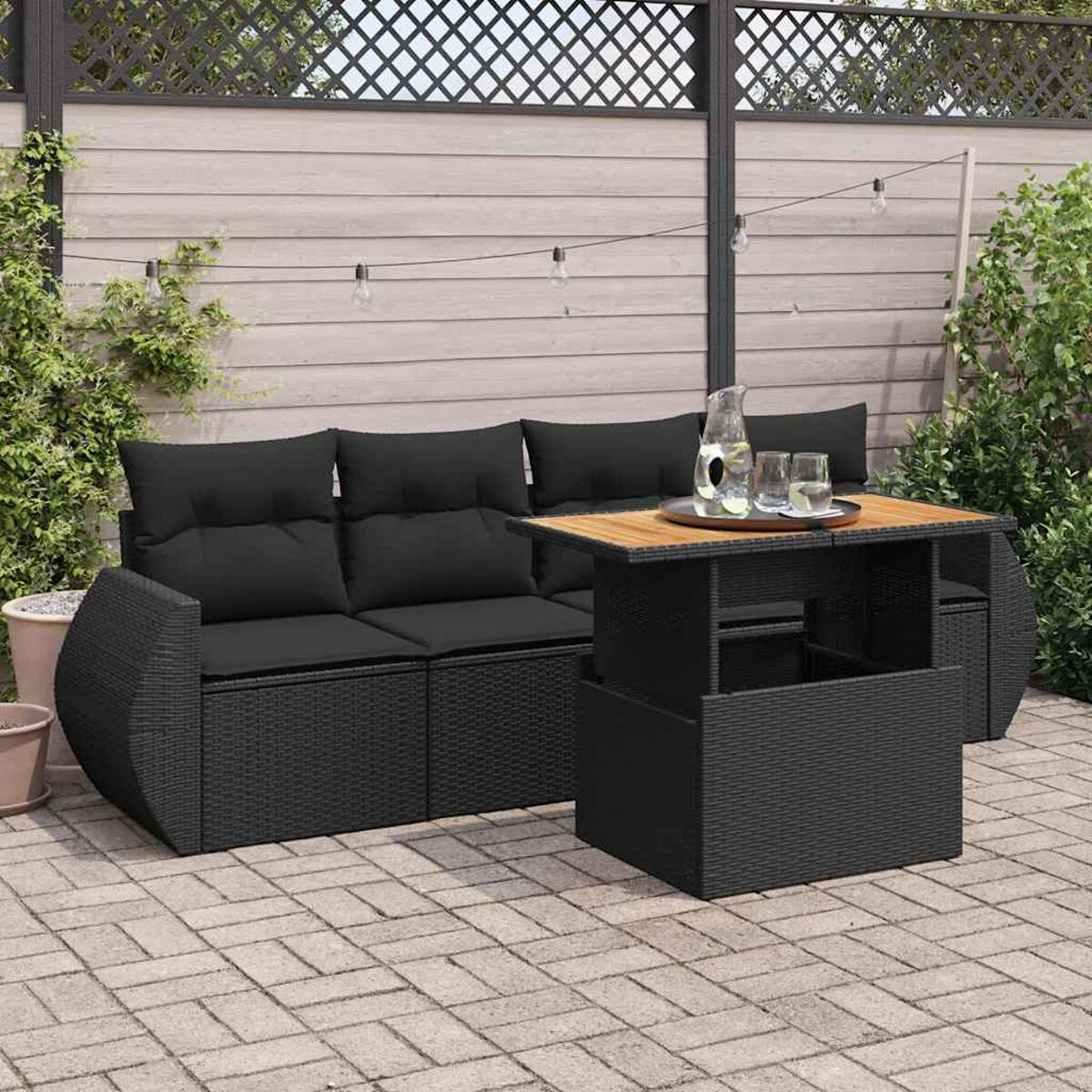 Set divano esterno in vimini nero con tavolo nero, su un patio in mattoni, con giardino sullo sfondo.