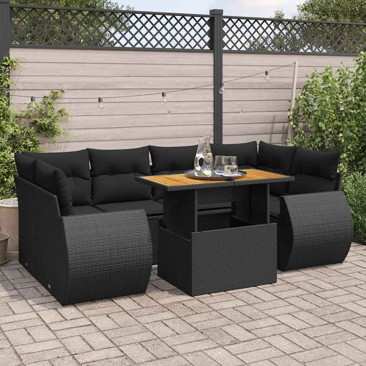 Set divano in rattan nero con un tavolo. Cuscini neri, tavolo con piano in legno, su un patio. Piante verdi e recinzione.