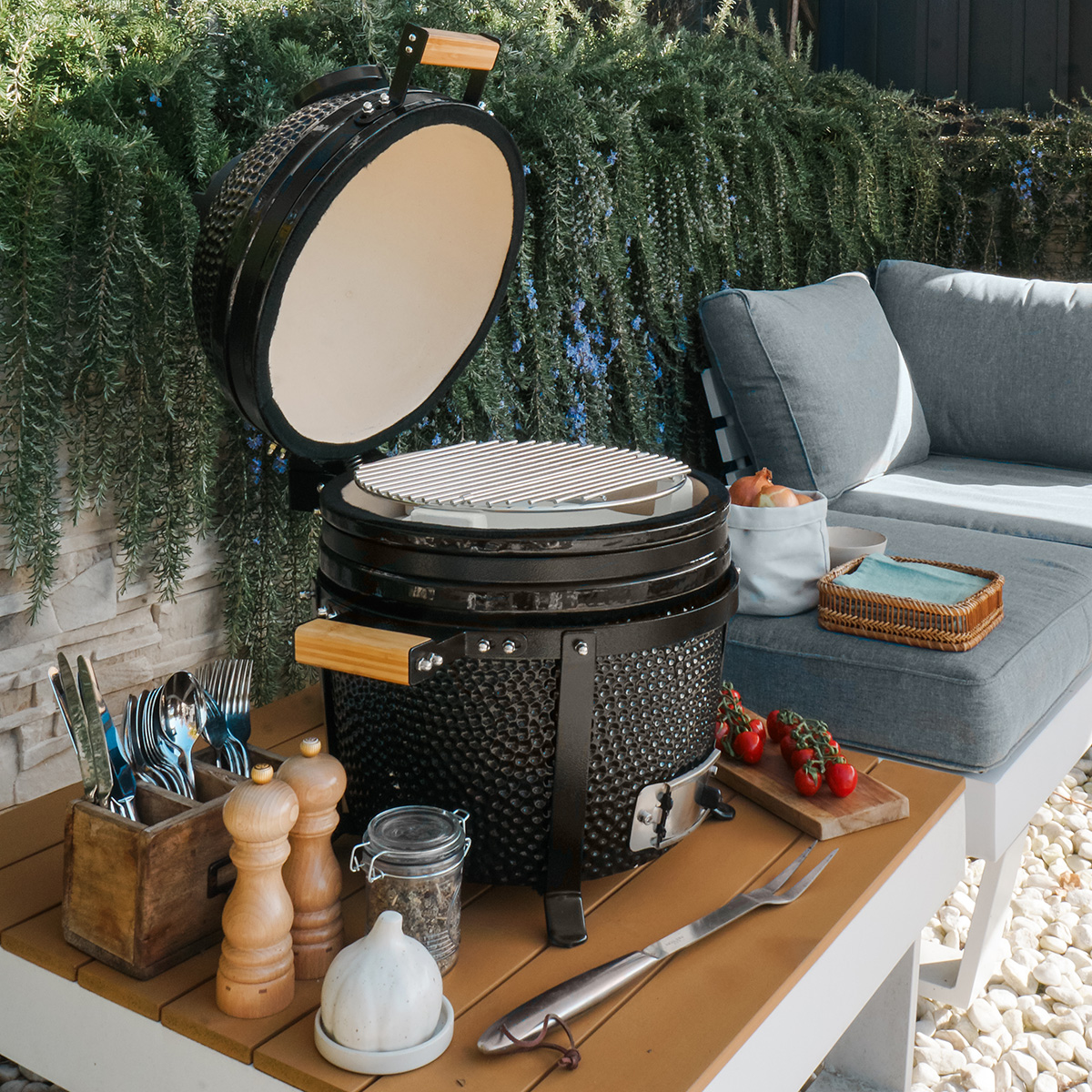 Outdoor scene with a black grill, food, and utensils on a wooden table. Green plants background.
