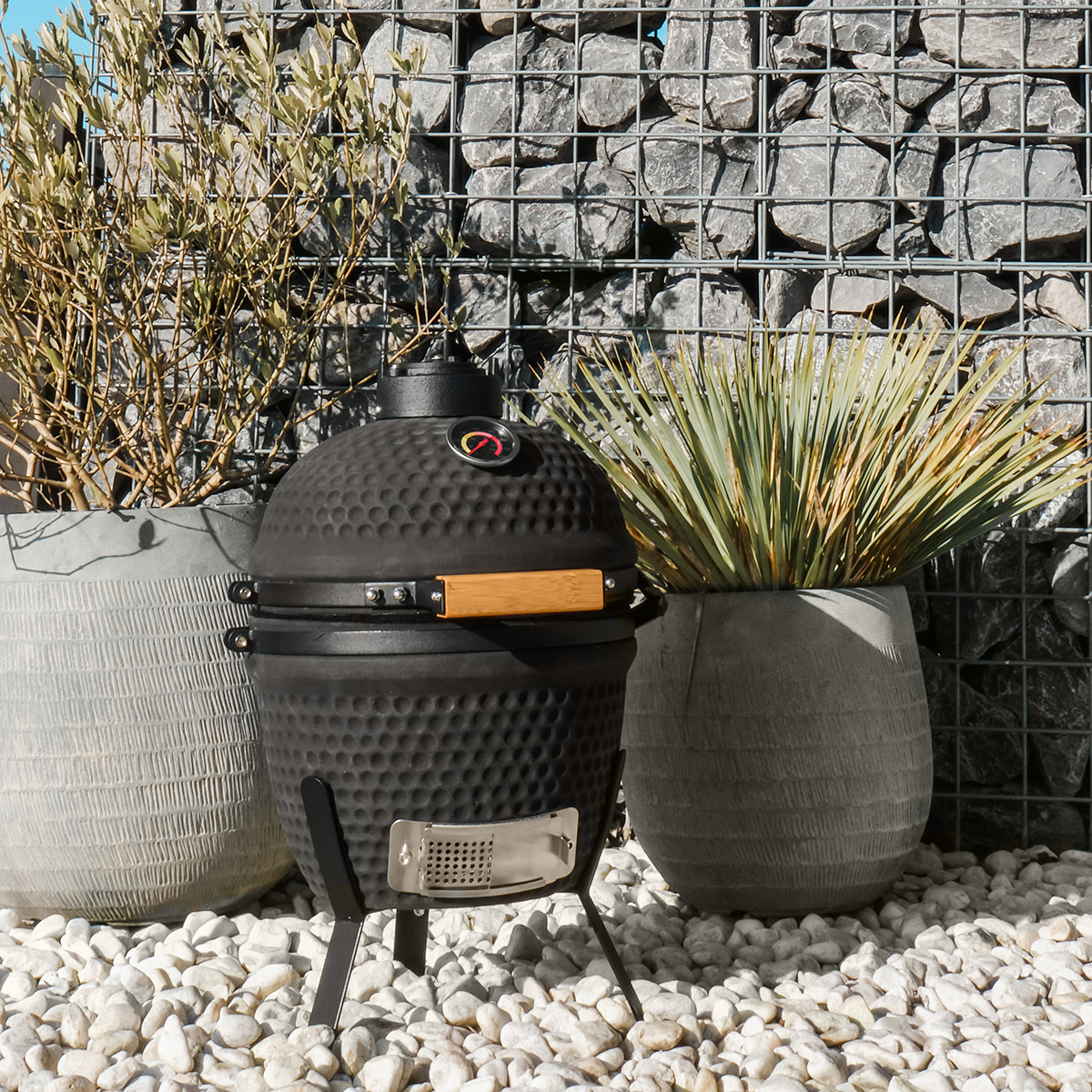 Ein schwarzer runder Smoker-Grill mit Griff und Thermometer, neben Topfpflanzen und einer Steinmauer.