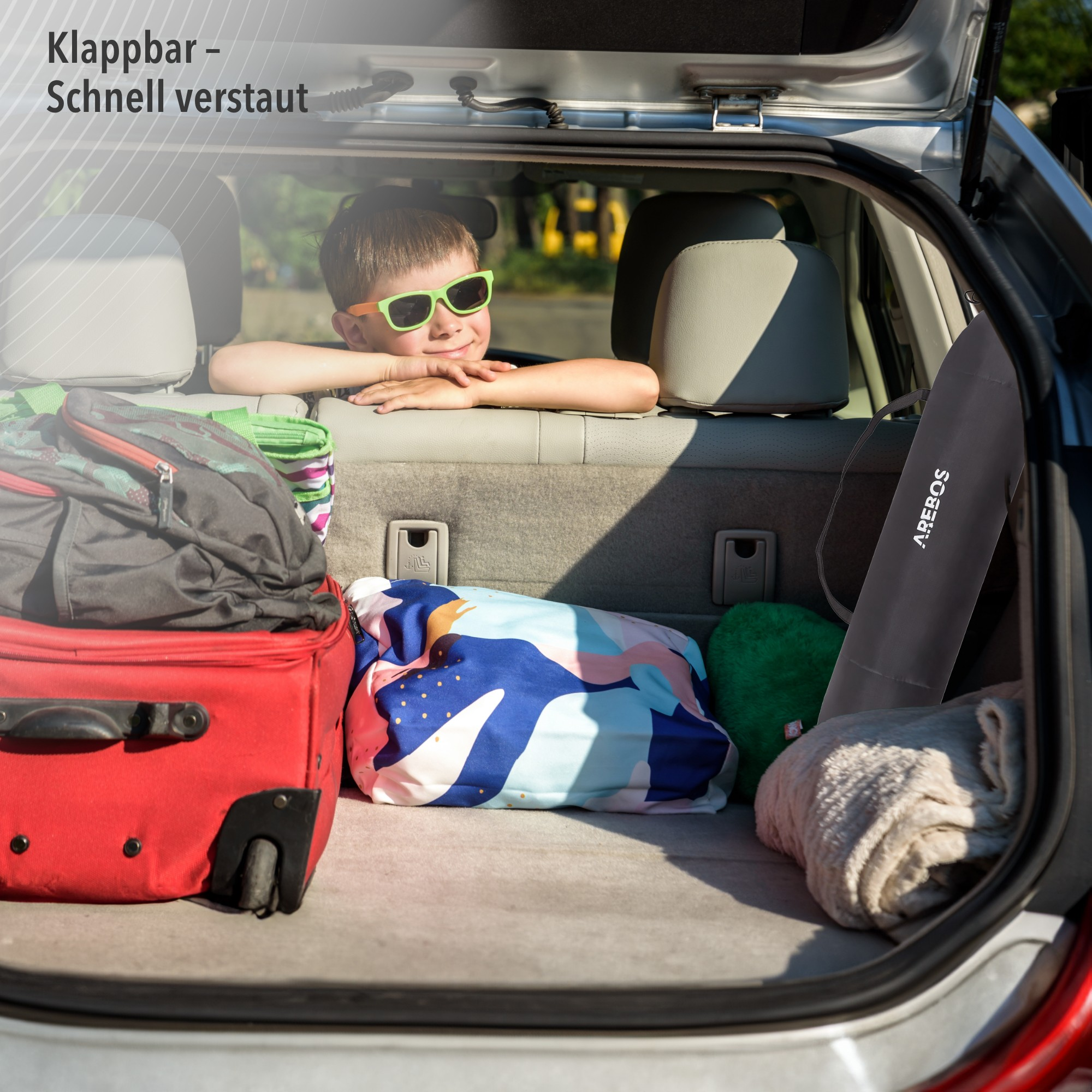 Ein Kind mit Sonnenbrille blickt aus einem Kofferraum voller Gepäck. Text: zusammenklappbar, schnell verstaut.