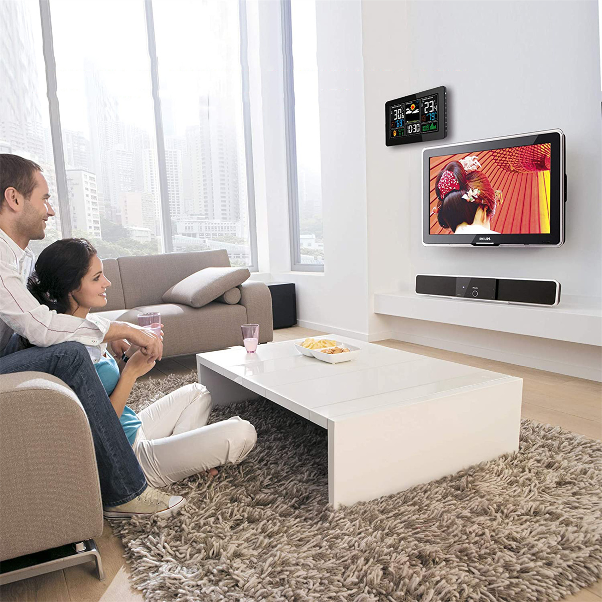 Ein Paar sitzt auf einem Sofa und schaut fern. An der Wand sind ein Fernseher und eine Wetterstation.