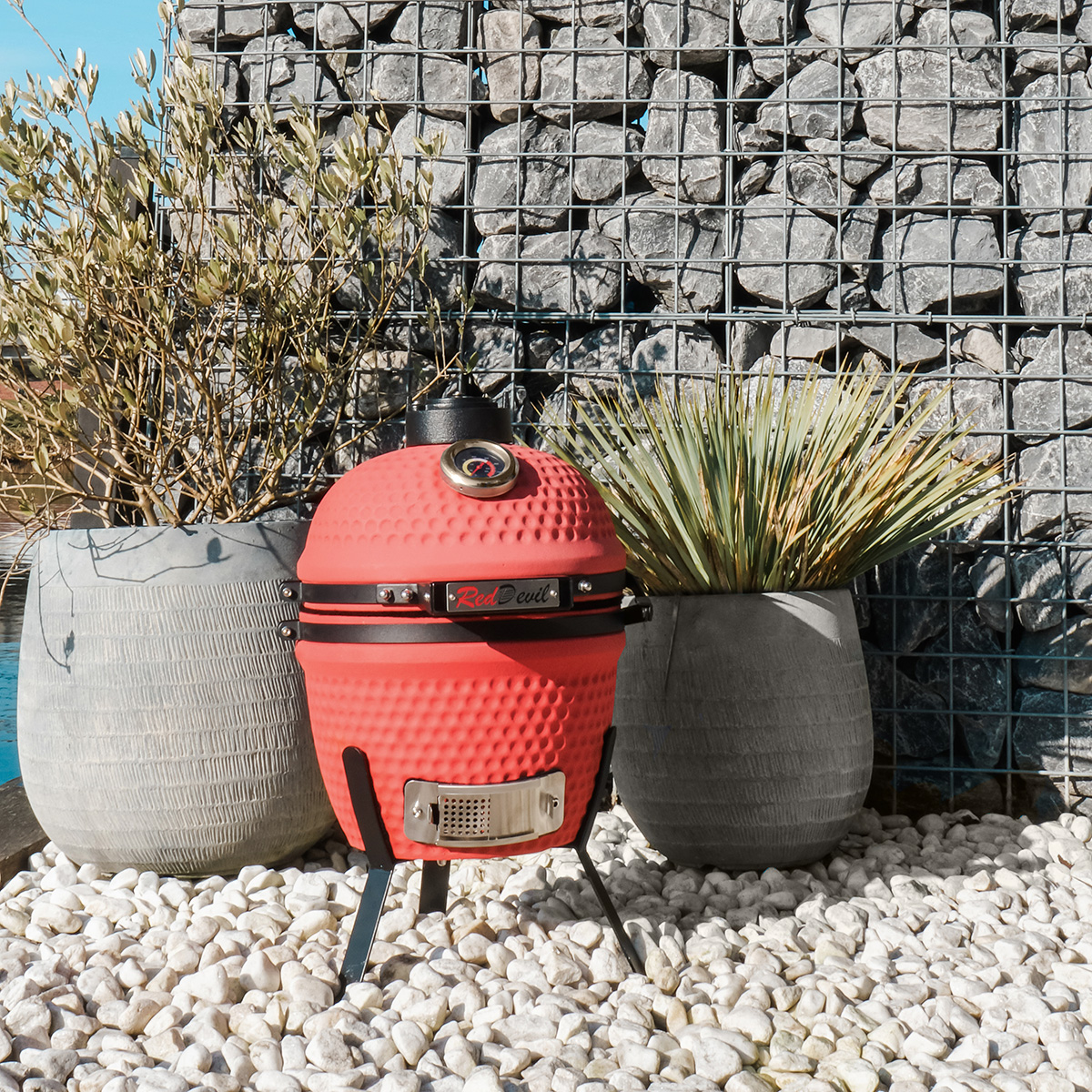 Roter Keramikgrill mit Thermometer, neben Pflanzen und einer Steinmauer.