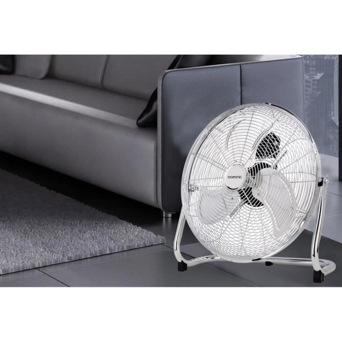 Silberner Ventilator. Der Ventilator steht auf einem hellgrauen Boden, im Hintergrund ein weißes Sofa.
