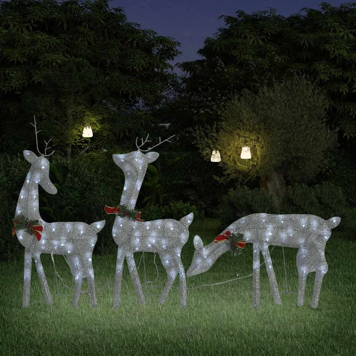 Drei beleuchtete Rentier-Weihnachtsdekorationen auf Gras, mit Bäumen und Lichtern im Hintergrund.