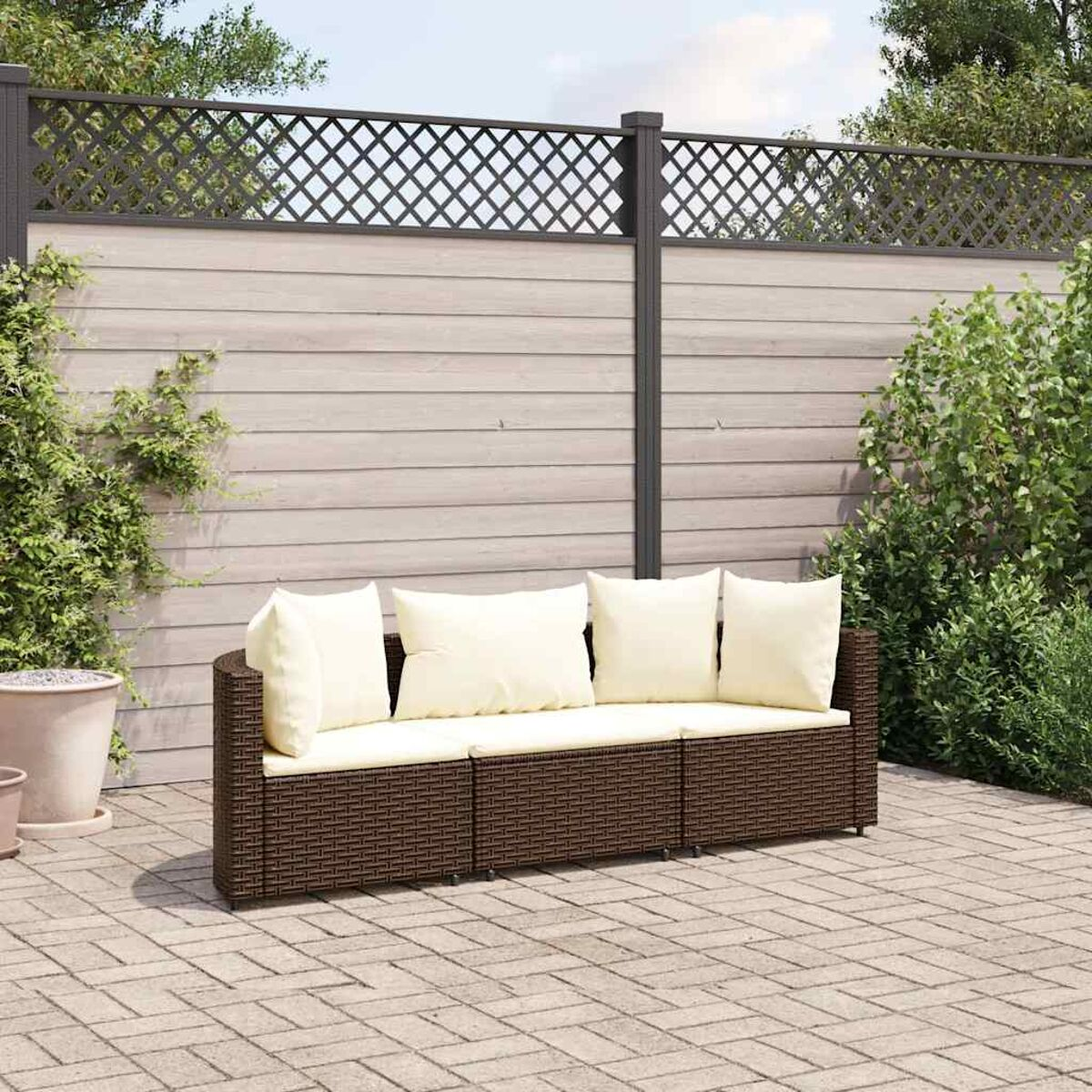 Divano in vimini marrone con cuscini color crema, in un giardino con recinzione e verde.