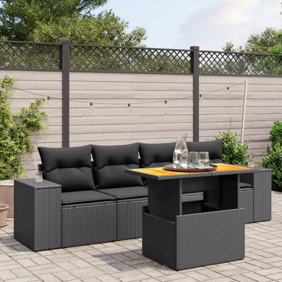 Outdoor-Rattan-Sofagarnitur mit Tisch und schwarzen Kissen. Auf Ziegelpflaster, mit Pflanzen und Zaun.