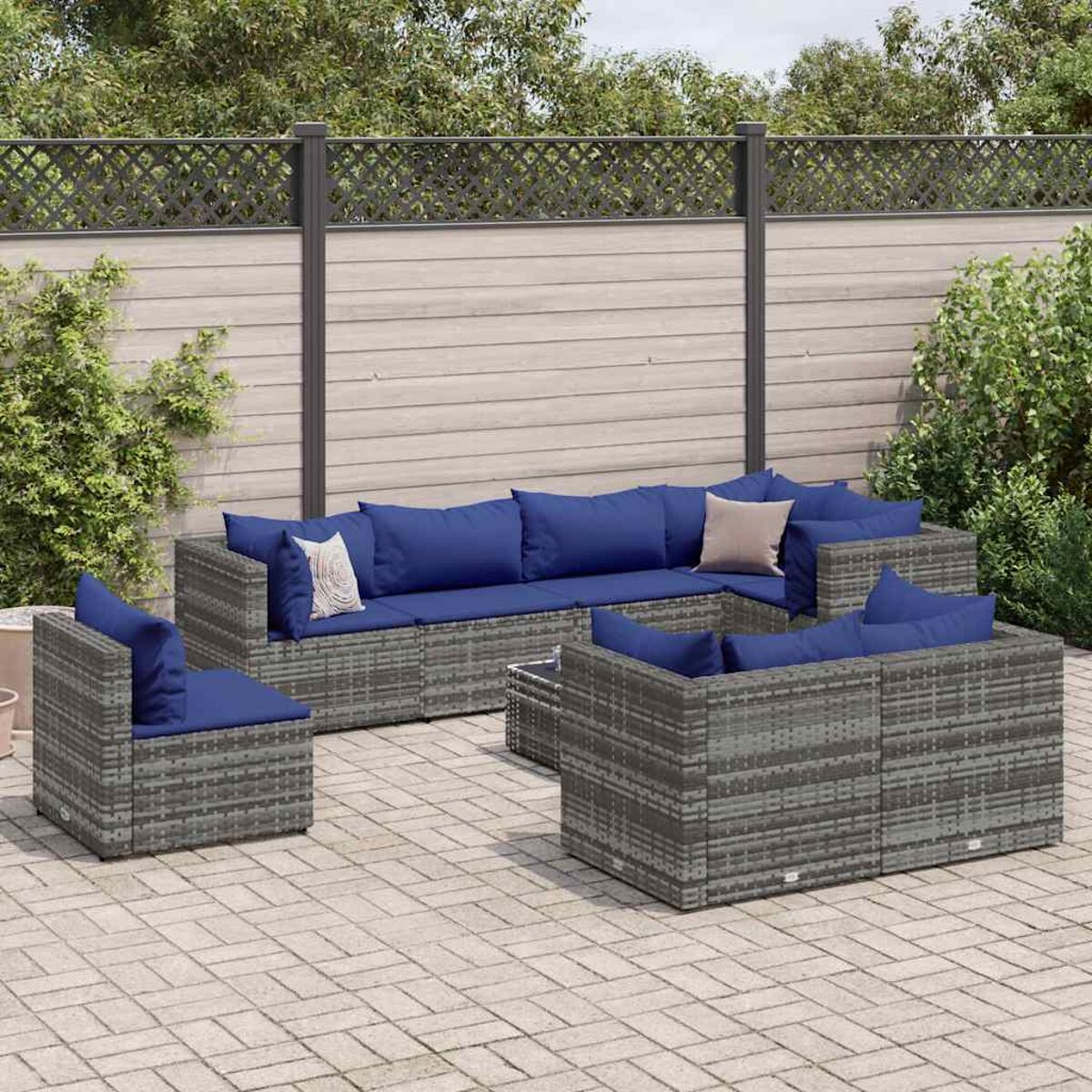 Outdoor-Sofagarnitur aus grauem Rattan mit blauen Kissen auf einer Backsteinterrasse. Hintergrund: Holzzaun und Pflanzen.